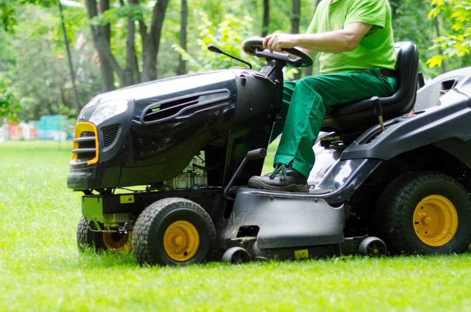 Tondeuse autoportée professionnelle verte et jaune en action sur une vaste pelouse de parc ou espace collectif entretenu, opérateur visible effectuant la tonte. Gazon vert uni, dégagé, sans obstacles. Scène en lumière naturelle de jour, paysagiste au travail.