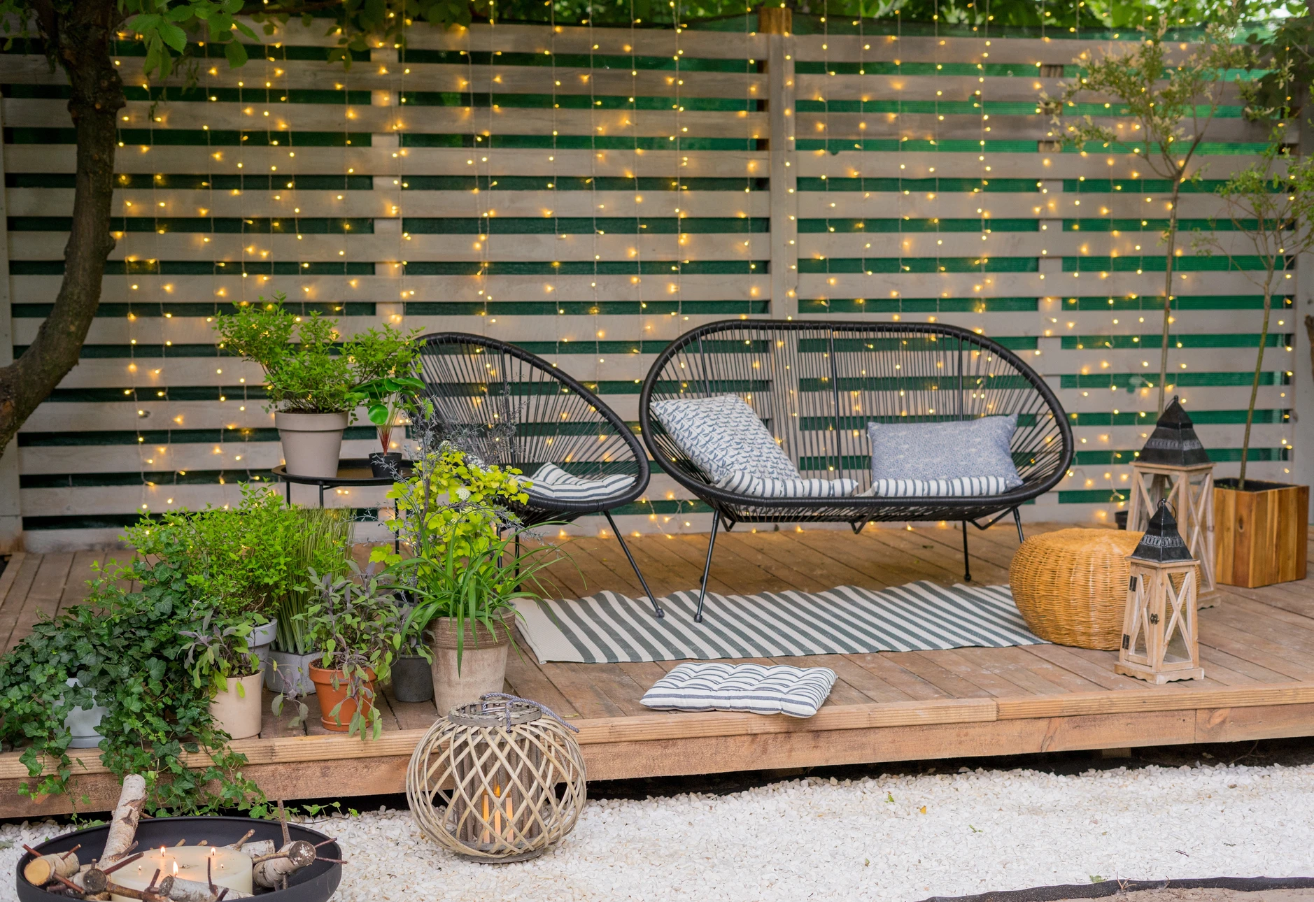 Paysagiste en tenue de travail taillant des arbustes en pot sur un balcon urbain en hauteur, avec vue sur les toits de la ville en arrière-plan. Le balcon est aménagé avec plusieurs bacs de plantation contenant des arbustes, du mobilier de jardin discret et des plantes bien entretenues. Le professionnel utilise un sécateur ou une cisaille de taille, concentré sur son ouvrage.